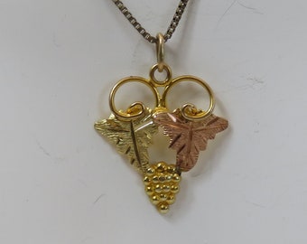 May include: A gold necklace with a pendant featuring a grapevine design with three leaves in gold, green, and rose gold. The pendant is in the shape of a heart.