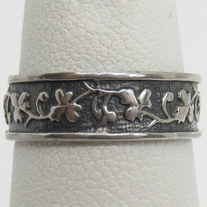 May include: A silver band ring with a textured finish and a delicate floral design. The band is wide and the design is intricate.