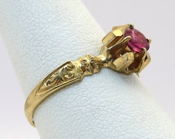 May include: A gold ring featuring a pink gemstone set in a prong setting. The band is adorned with an intricate floral design.