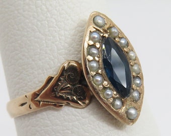 Op de afbeelding: Een goudkleurige ring met een donkerblauwe, marquise-vormige edelsteen, omringd door kleine parels. De ringband heeft een sierlijk ontwerp met geëtste details, wat een vintage uitstraling geeft.