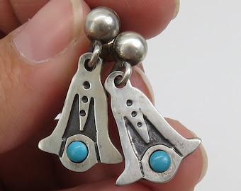 May include: A pair of silver dangle earrings with turquoise stones. The earrings are in the shape of a bell with a geometric design.