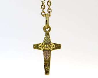 Pode incluir: Um colar com um pingente de cruz de ouro com um pequeno design de folha verde e marrom e contas douradas. O colar está sobre um fundo branco.