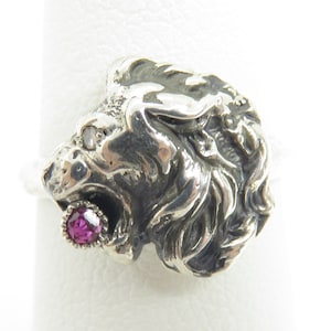 May include: A silver ring with a detailed lion head design. The lion's mouth is open and a pink gemstone is set in its mouth.