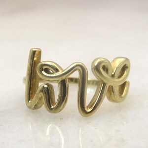 Peut inclure: Une bague dorée épelant le mot "love" dans une police cursive. La bague est faite d'un matériau métallique brillant et repose sur une surface réfléchissante. Les lettres sont reliées et forment un motif continu.