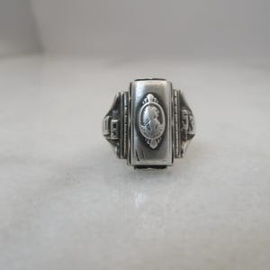 May include: A silver-toned ring with a rectangular face featuring a cameo-style design. The ring has decorative side detailing and the year 1953 is visible on the sides. The ring is likely a class ring or commemorative piece.