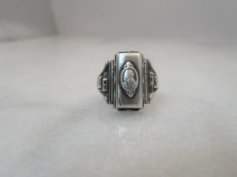 May include: A silver-toned ring with a rectangular face featuring a cameo-style design. The ring has decorative side detailing and the year 1953 is visible on the sides. The ring is likely a class ring or commemorative piece.