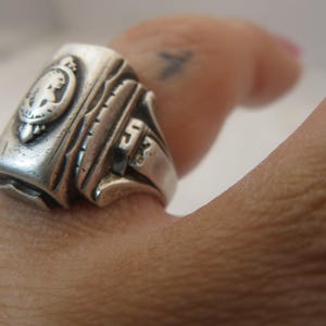 May include: Close-up of a silver ring with a detailed design. The ring features a crest with a profile and the number "53". The ring is worn on a finger.