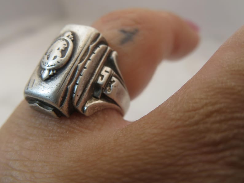 May include: Close-up of a silver ring with a detailed design. The ring features a crest with a profile and the number "53". The ring is worn on a finger.