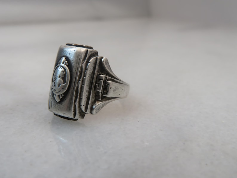 May include: A silver-toned ring with a rectangular face featuring a detailed relief of a woman's profile within a circular frame. The ring has a textured band and a classic, vintage design.
