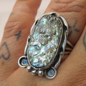 May include: A silver ring with a large oval iridescent abalone shell set in a silver bezel. The ring has a decorative silver band with a textured design.