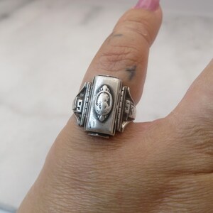 May include: A silver-toned class ring with a rectangular face featuring a cameo design. The ring has the numbers "19" and "53" on either side of the face. The ring is worn on a finger.