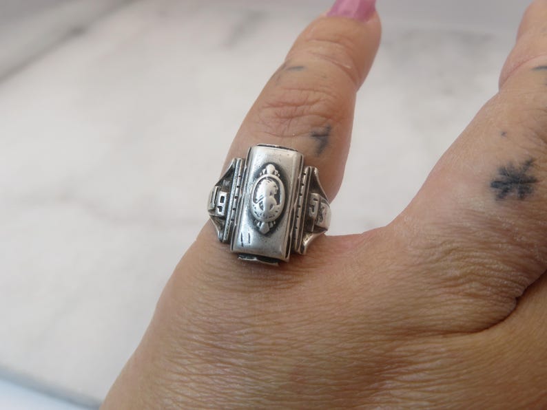 May include: A silver-toned class ring with a rectangular face featuring a cameo design. The ring has the numbers "19" and "53" on either side of the face. The ring is worn on a finger.