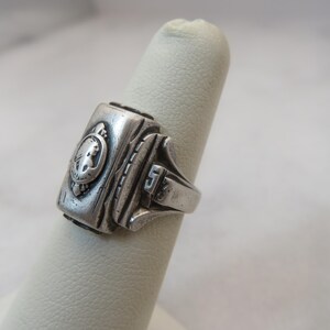 May include: A silver-toned class ring with a rectangular face featuring a relief of a person's profile within a circular emblem. The ring has a detailed design with the number "5" on the side.