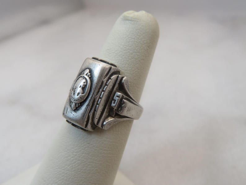 May include: A silver-toned class ring with a rectangular face featuring a relief of a person's profile within a circular emblem. The ring has a detailed design with the number "5" on the side.