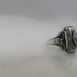 May include: A silver-toned ring with a rectangular face and ornate detailing. The ring features a detailed design on the face, with a small circular emblem. The band has a classic, slightly worn appearance.