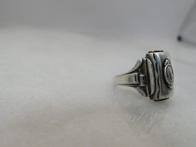 May include: A silver-toned ring with a rectangular face and ornate detailing. The ring features a detailed design on the face, with a small circular emblem. The band has a classic, slightly worn appearance.