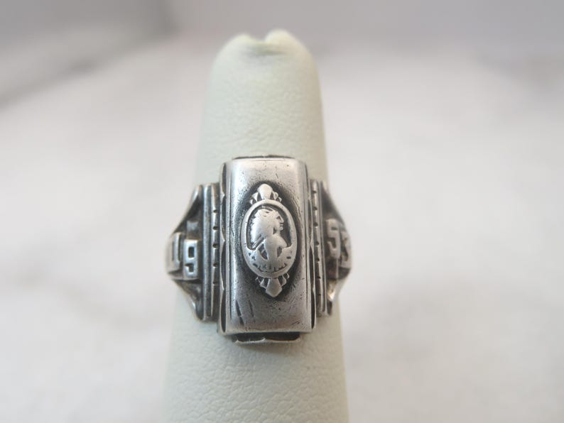 May include: Silver-toned class ring with a rectangular face featuring a cameo-style design and the year 1953. The ring is displayed on a finger, showcasing its intricate details and vintage aesthetic.