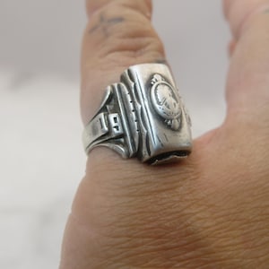 May include: A silver-toned ring with a rectangular face, featuring a crest design and the number "19". The ring is worn on a finger, showcasing its vintage style and detailed craftsmanship. The ring's design includes a detailed crest and decorative elements.
