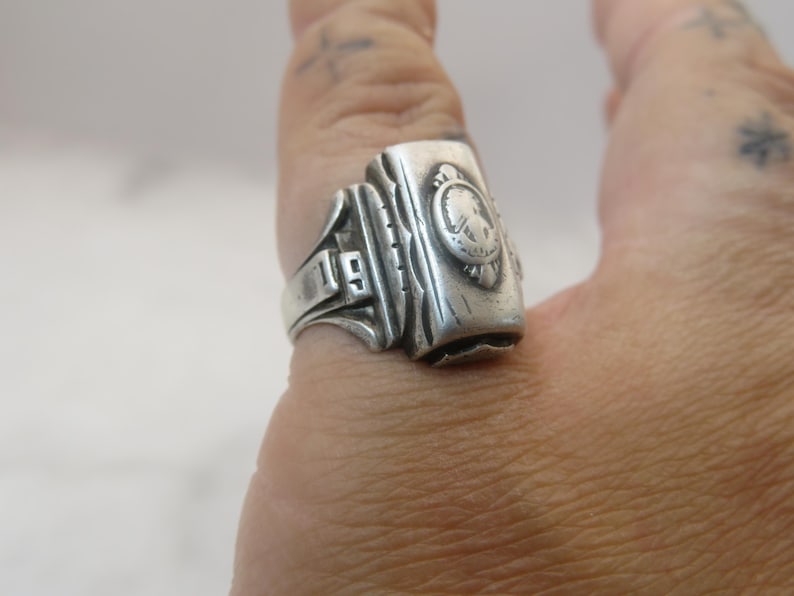 May include: A silver-toned ring with a rectangular face, featuring a crest design and the number "19". The ring is worn on a finger, showcasing its vintage style and detailed craftsmanship. The ring's design includes a detailed crest and decorative elements.