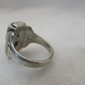 May include: A silver ring with a rectangular, textured face. The ring's band is smooth and rounded, and the face has a detailed, possibly engraved design. The ring is set against a neutral background, highlighting its metallic sheen.