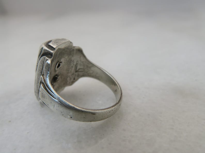 May include: A silver ring with a rectangular, textured face. The ring's band is smooth and rounded, and the face has a detailed, possibly engraved design. The ring is set against a neutral background, highlighting its metallic sheen.