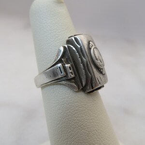 May include: A silver-toned ring with a rectangular face and a detailed design. The ring features the number "19" on the band and a circular emblem on the face. The face has a textured, wood-like appearance.