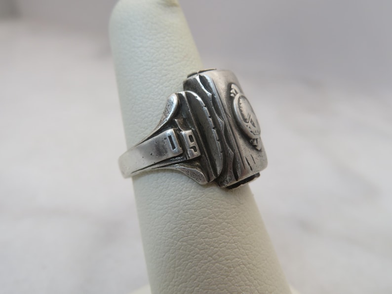 May include: A silver-toned ring with a rectangular face and a detailed design. The ring features the number "19" on the band and a circular emblem on the face. The face has a textured, wood-like appearance.