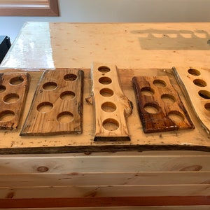 May include: Five wooden beer flight boards with circular cutouts for glasses. The boards vary in shape and wood grain, with a natural, rustic aesthetic. Some boards have a dark, glossy finish, while others are lighter.