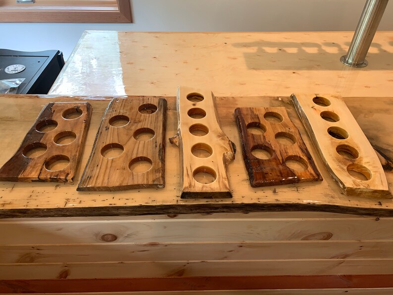 May include: Five wooden beer flight boards with circular cutouts for glasses. The boards vary in shape and wood grain, with a natural, rustic aesthetic. Some boards have a dark, glossy finish, while others are lighter.