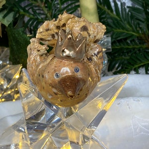 May include: A handcrafted ceramic bird figurine with a gold crown, resting on a clear, faceted star. The bird has a speckled, tan body, blue eyes, and a detailed beak. The star is made of clear glass and reflects light, creating a festive display.