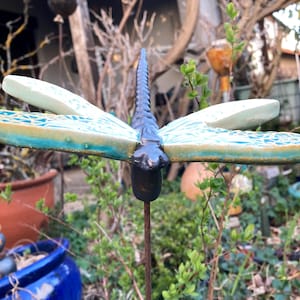 Könnte beinhalten: Ein dekorativer Gartenstecker in Form einer Libelle. Die Libelle hat einen dunkelblauen Körper und Kopf mit hellblauen und weißen Flügeln. Die Flügel haben ein strukturiertes Design. Der Stecker ist aus Metall und steht in einem Garten.
