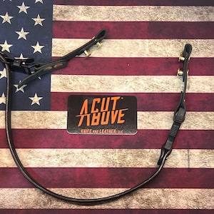 Könnte beinhalten: Ein schwarzes Lederband mit Messingbeschlägen und einer schwarzen Kunststoffschnalle. Das Band liegt auf einem rot-weiß-blauen amerikanischen Flaggenhintergrund. Der Text "A CUT ABOVE" befindet sich auf einem schwarzen Aufkleber mit orangefarbenem Text.