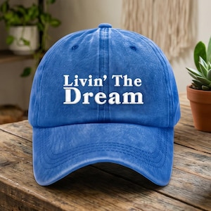 May include: A blue baseball cap with a distressed finish featuring the white embroidered text Livin' The Dream on the front panel. The cap has a curved brim and a structured crown with visible stitching details across the fabric panels.