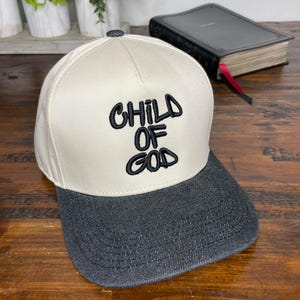 Könnte beinhalten: Eine beige und schwarze Baseballkappe mit den Worten "CHILD OF GOD" in Schwarz auf der Vorderseite gestickt.