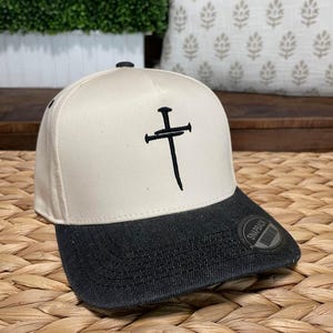 Könnte beinhalten: Eine weiße Baseballkappe mit einem schwarzen Kreuzdesign und einem schwarzen Schirm. Der Schirm hat ein schwarzes "Snapback"-Etikett.