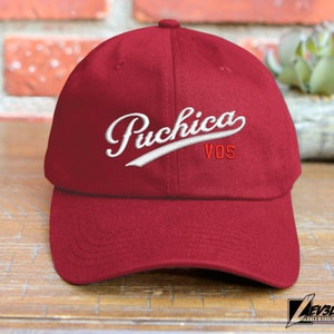 May include: A red baseball cap with a white embroidered logo that says "Puchica VOS".