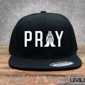 May include: Black and white baseball cap with the word "PRAY" in white lettering and a pair of hands in a praying position.