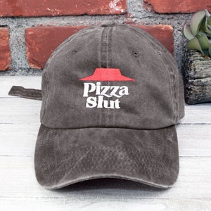 May include: A dark gray baseball cap with a curved brim. The cap features the embroidered text "Pizza Slut" in white, with a red logo above the text. The cap is made of a soft, worn material.