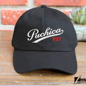 May include: Black baseball cap with white embroidered text "Puchica VOS" in a cursive font.