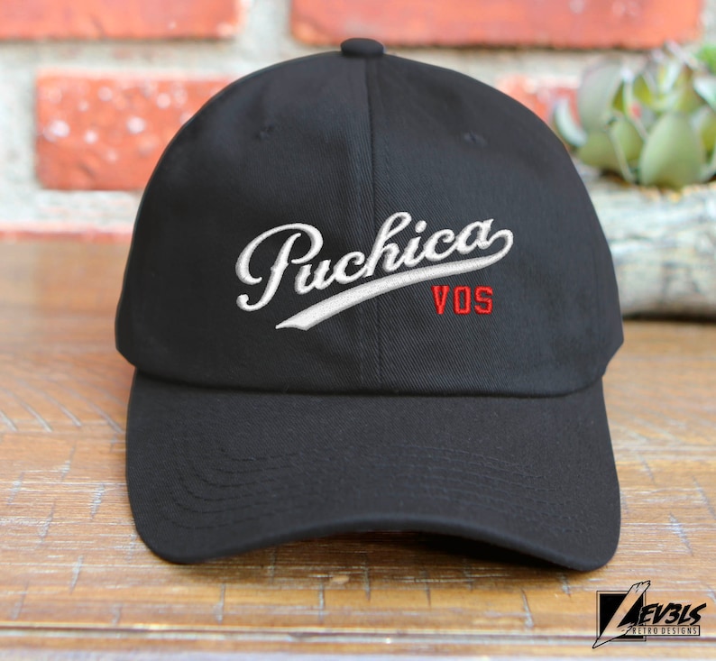 May include: Black baseball cap with white embroidered text "Puchica VOS" in a cursive font.