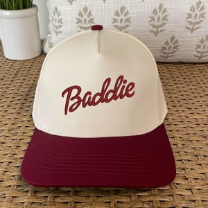 May include: A white and burgundy baseball cap with the word "Baddie" embroidered in red on the front.