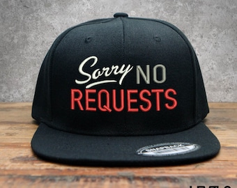 以下が含まれることがあります： 黒のベースボールキャップ。前面に白の刺繍で「Sorry No Requests」と入っています。キャップは黒のスナップバックです。