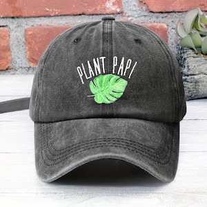 May include: A black, distressed, adjustable baseball cap with the text "PLANT PAPI" embroidered in white and a green monstera leaf embroidered below the text.
