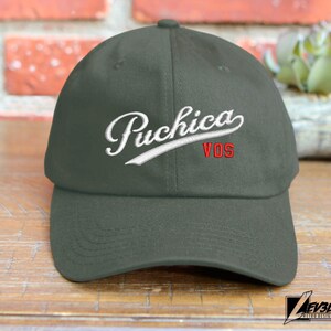 May include: A dark green baseball cap with a white embroidered logo that says "Puchica VOS" in a cursive font.