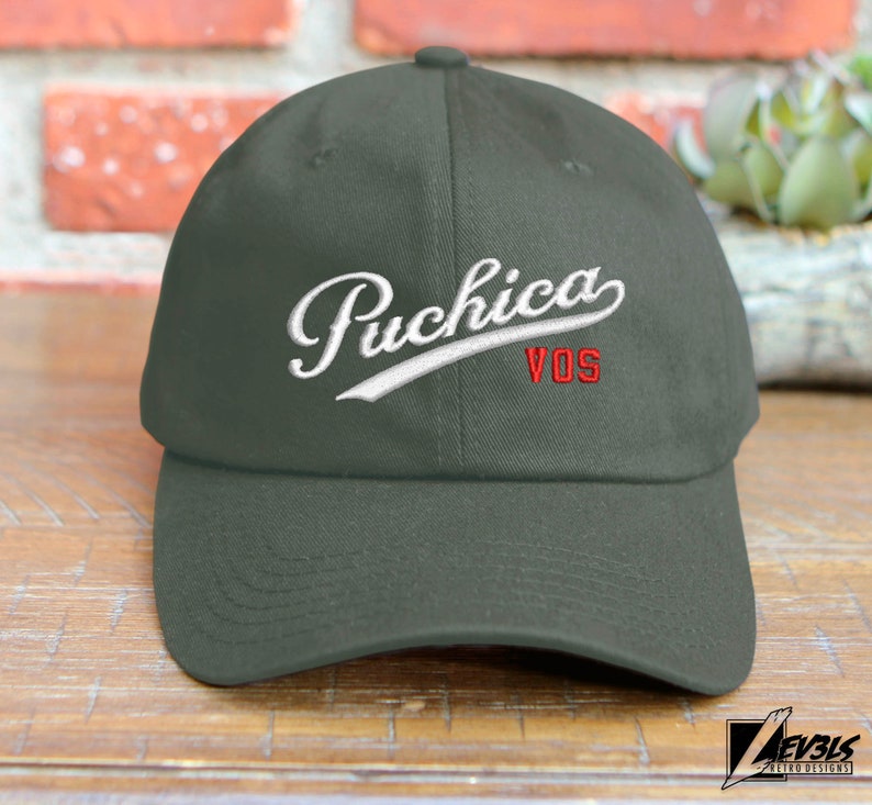 May include: A dark green baseball cap with a white embroidered logo that says "Puchica VOS" in a cursive font.