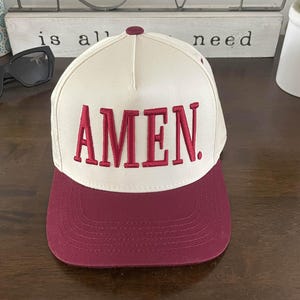 以下が含まれることがあります： クリーム色のクラウンとマルーンのつばが付いた野球帽。「AMEN」の文字がマルーンのブロック体で前面に刺繍されています。帽子はカーブしたつばと上部にボタンが付いています。