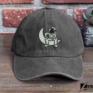 May include: A dark gray, distressed, cotton baseball cap with an embroidered design of an astronaut sitting on a crescent moon.