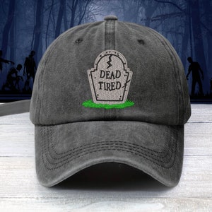 May include: A black, distressed, cotton baseball cap with an embroidered image of a tombstone that reads "Dead Tired".