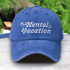 以下が含まれることがあります： 白い筆記体で「Mental Vacation」と刺繍された青い野球帽。つばがカーブしており、調節可能なストラップが付いています。デザインにはヤシの木と2羽の鳥が含まれています。