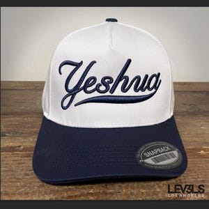 May include: White and navy blue baseball cap with the word "Yeshua" embroidered in navy blue on the front. The cap has a snap closure and a label that says "Snapback" and "High Quality" on the back.